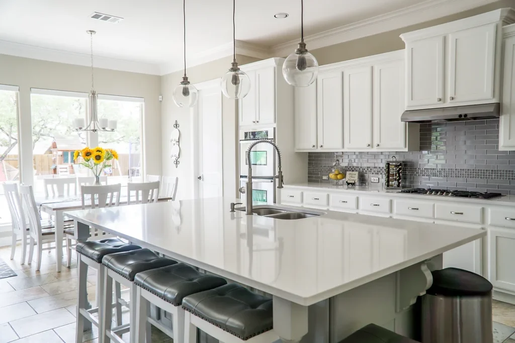 A newly remodeled kitchen with a large island with a white quartz countertop surrounded by white cabinets with a gray tile backsplash